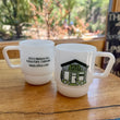 Load image into Gallery viewer, Two white ceramic coffee mugs with built-in handles sit on a wooden table in front of a window. One mug has black printed text reading '470 E Elkhorn Ave Estes Park, Colorado kindcoffee.
